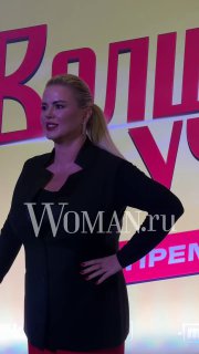 Анна Семенович стоит в полный рост на сцене или фотоколле: чёрный костюм, поза для фото, на заднике крупные буквы и логотип мероприятия с водяным знаком Woman.ru