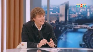 Кадр студии утреннего шоу: мужчина в роли ведущего сидит за столом на фоне панорамного окна с видом на город, логотип ТНТ в углу.