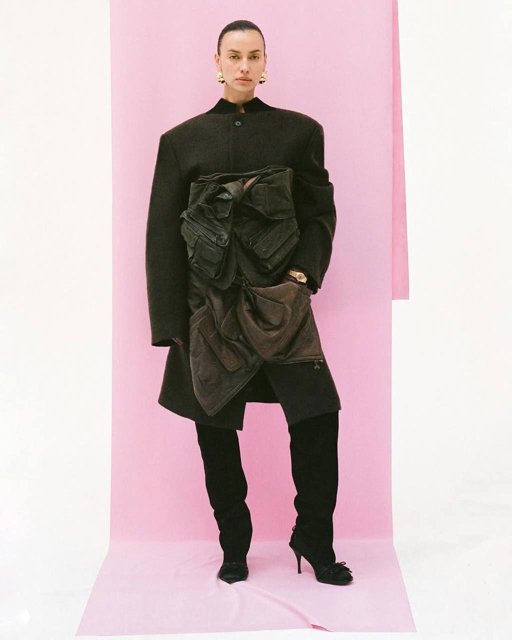 Полнотелый кадр Ирины Шейк на розовом фоне в строгом пальто и высоких ботфортах — образ в духе высокой моды.