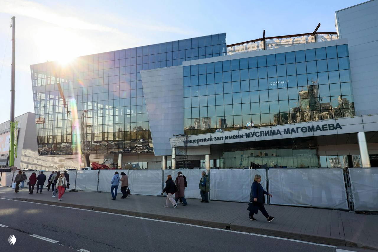 Дневное фото фасада концертного зала «Крокус»: ограждение у входа, люди проходят мимо, видны следы ремонта и охраны территории