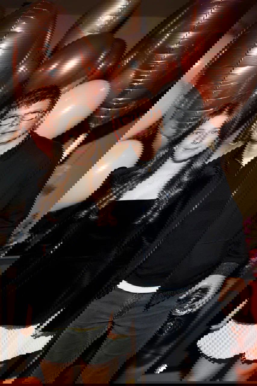 Девушка-подросток и взрослый мужчина позируют рядом друг с другом на фоне золотисто-розовых и белых воздушных шаров во время семейного торжества.