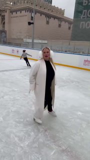 Анна Семенович в длинном светлом пальто стоит на льду городского катка в полный рост; вокруг видны бортики и другие фигуристы.