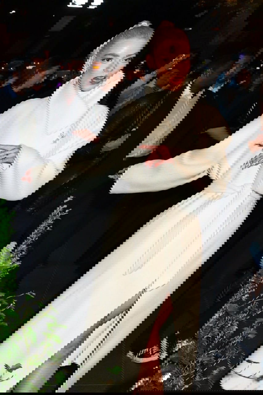 Аня Тейлор‑Джой вечером в элегантном белом длинном пальто с поясом, на высоких каблуках, окружение — фотографы и прохожие.