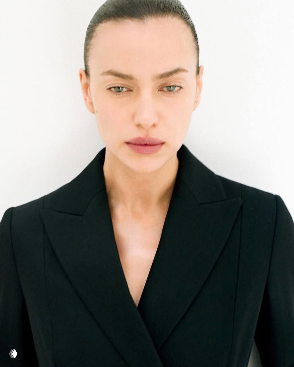 Крупный строгий портрет Ирины Шейк в чёрном пиджаке на белом фоне — акцент на чертах лица и минималистичном стиле.