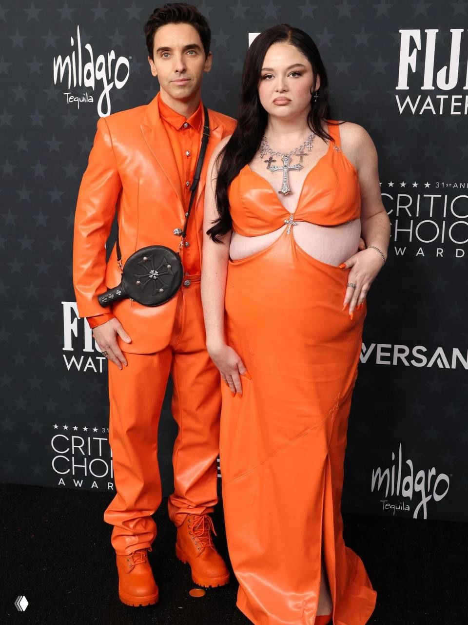 Пара на ковровой дорожке в ярко‑оранжевых кожаных нарядах; образ явно отсылает к мемному выходу с премьеры «Марти Суприм», фон с логотипами мероприятия.