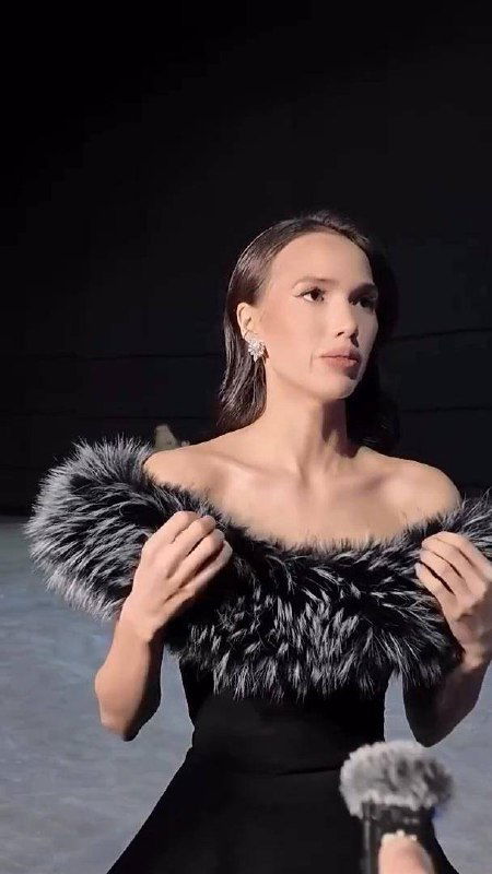 Женщина в чёрном вечернем платье с меховой отделкой позирует на сцене; видны плечи, серьги и микрофон внизу кадра.