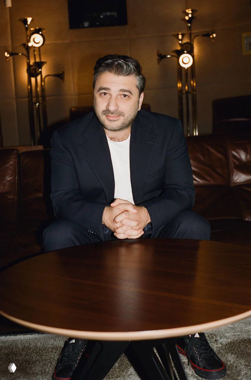 Мужской портрет в тёплом интерьере: мужчина в тёмном пиджаке сидит за круглым столом, мягкое освещение и атмосферная кинематографичная съёмка.