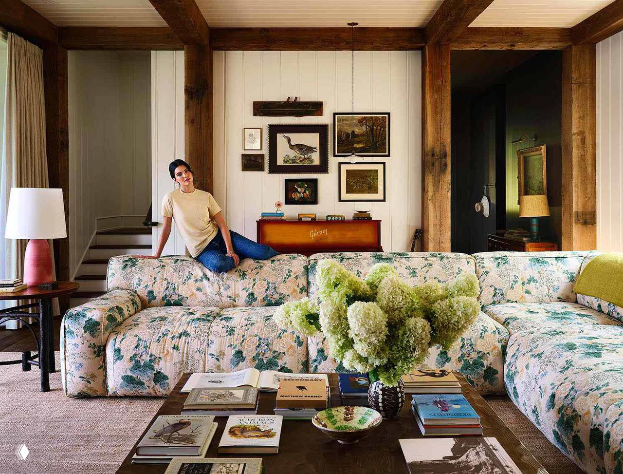 Гостиная с большим цветочным диваном, журнальным столиком с книгами и Кендалл, сидящей на диване — сочетание винтажа и мягких текстур в интерьере.