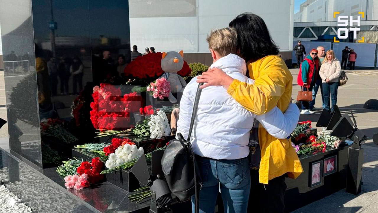 Люди у мемориала: две женщины обнимаются рядом с цветами, игрушками и венками, скорбный момент памяти о погибших в атаке