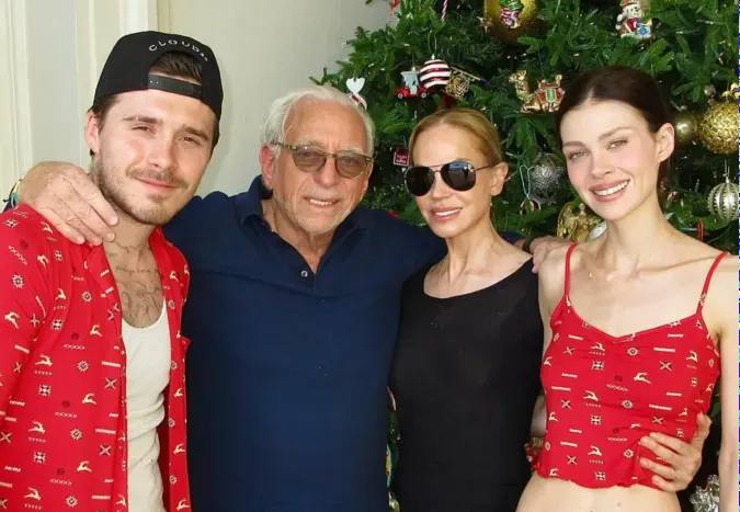 Никола Пельтц и Бруклин рядом с пожилой парой у украшенной рождественской ёлки: тёплый семейный портрет в домашней атмосфере.