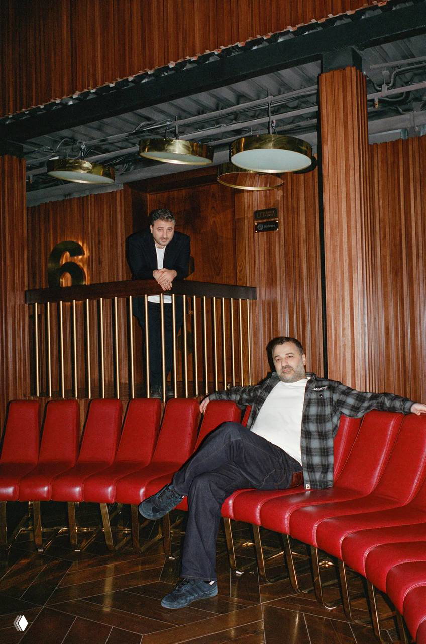 Два мужчины в кинозале: один опирается на балюстраду, другой расслабленно сидит в красных креслах; винтажная обстановка и тёплые тона.