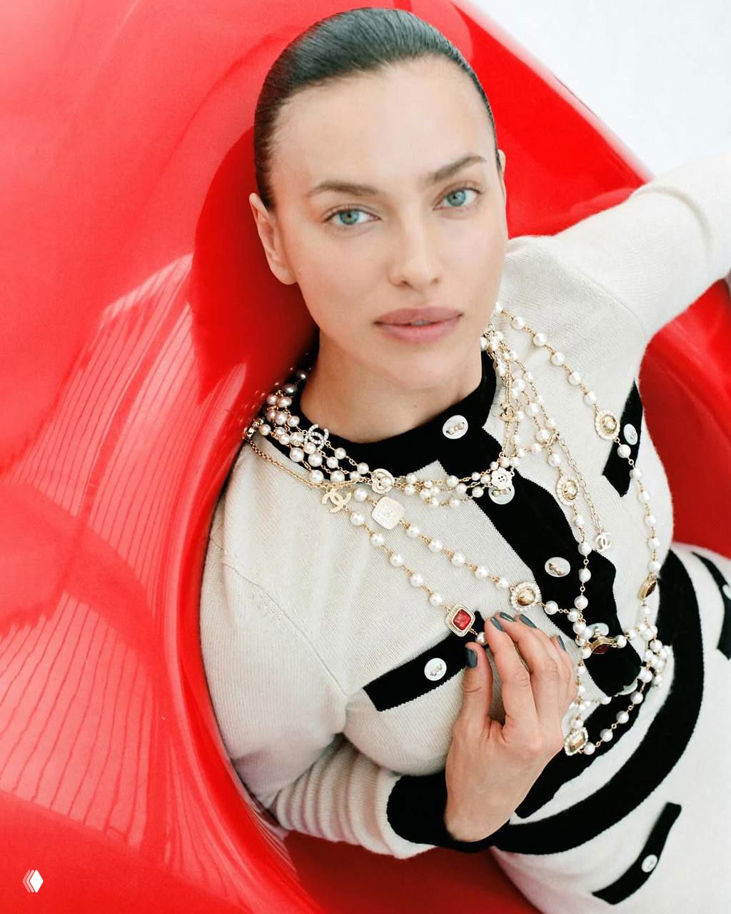 Крупный план Ирины Шейк в светлом наряде с многослойными бусами на ярком красном фоне — акцент на украшениях и лице модели.