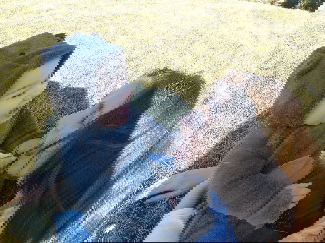 Крупный план: улыбающийся малыш Мерлин в тёплом комбинезоне и радостная Наталья Орейро, они лежат на солнце на траве.