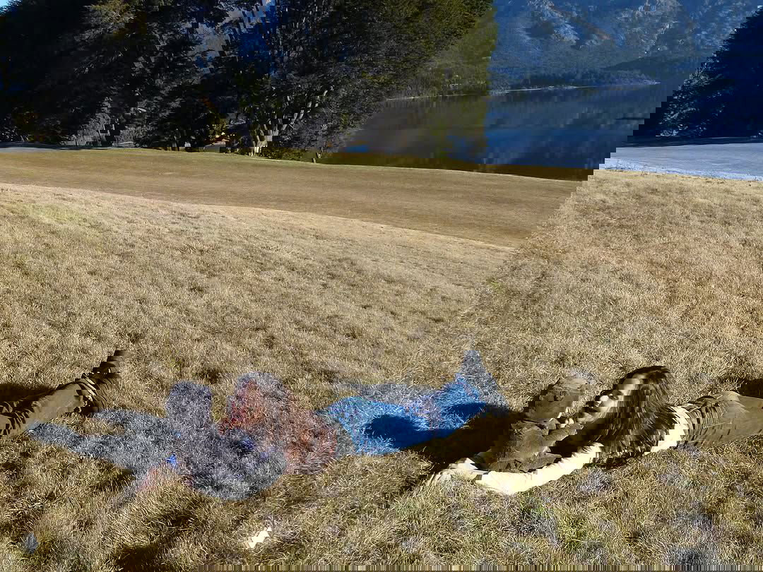 Широкий план: Наталья Орейро лежит на лугу рядом с ребёнком, просторный вид на поле, озеро и горы — семейная фотосессия.