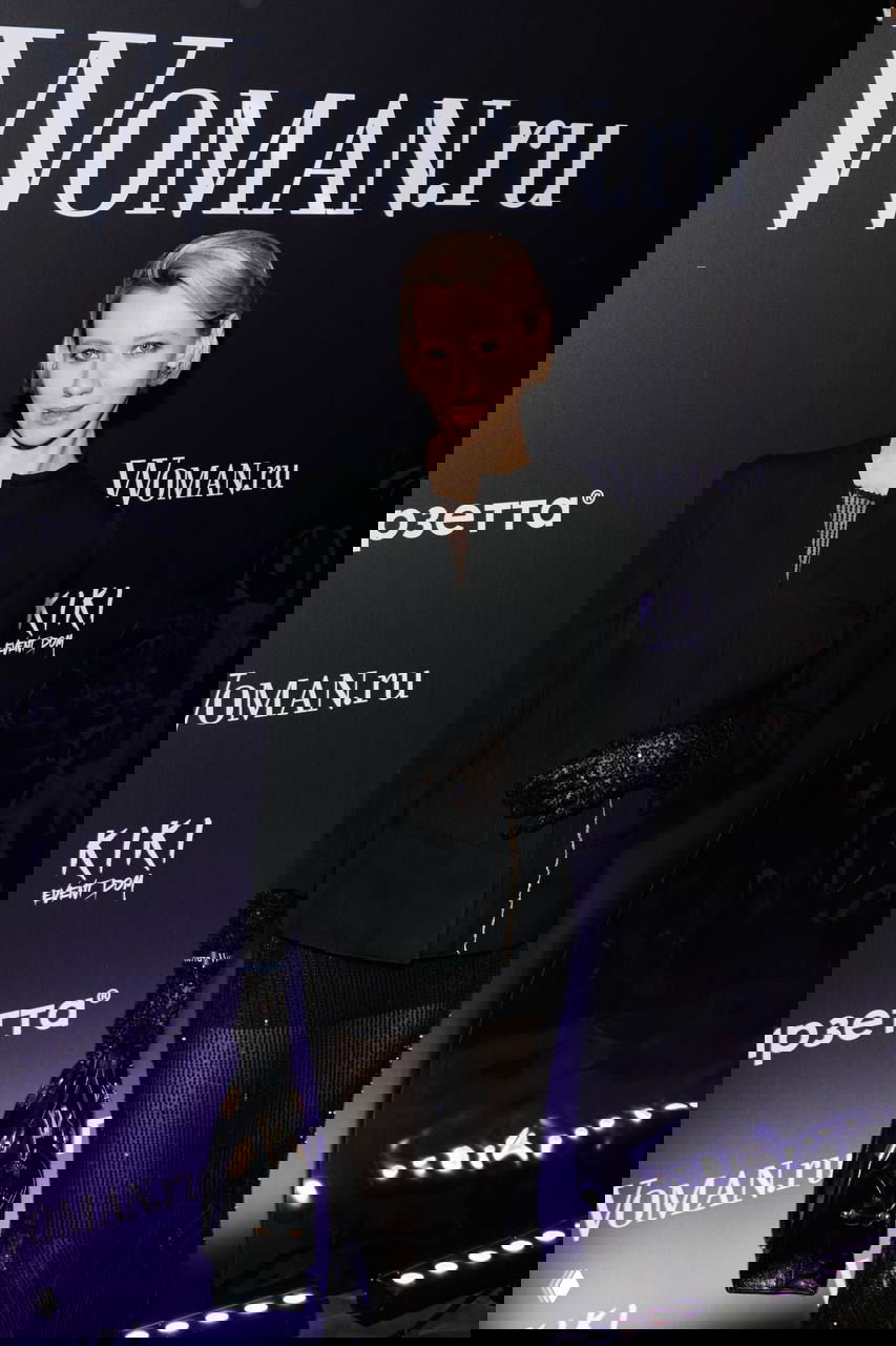 Гостья в элегантном черном образе с перчатками и минимальным украшением позирует на фоне логотипа Woman.ru на мероприятии.