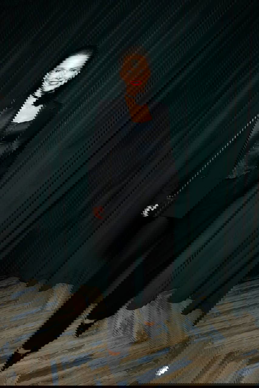 Женщина в чёрном костюме с корсетом и сумкой стоит в полный рост на фоне зелёной шторы, спокойная улыбка и вечерний образ.