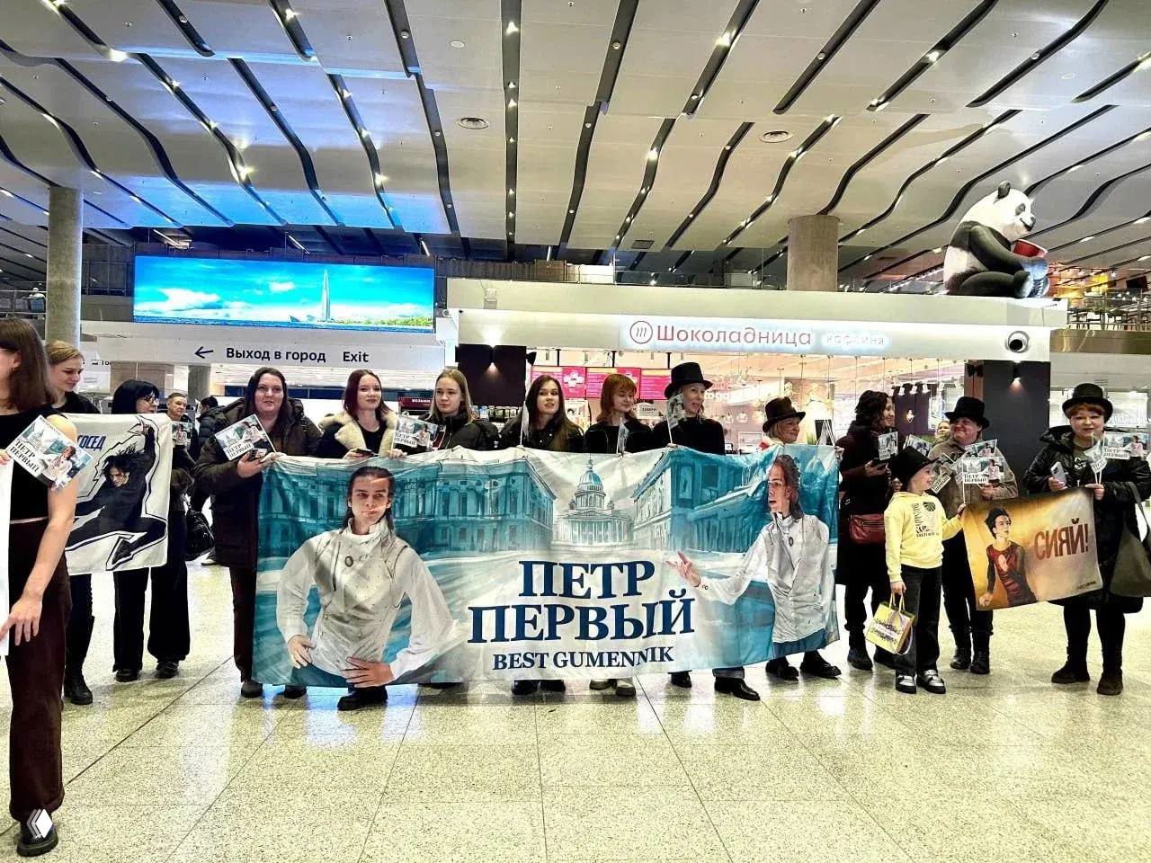 Группа поклонников в холле аэропорта Пулково держит большой баннер с надписью «Петр Первый», плакаты и журналы в ожидании фигуриста