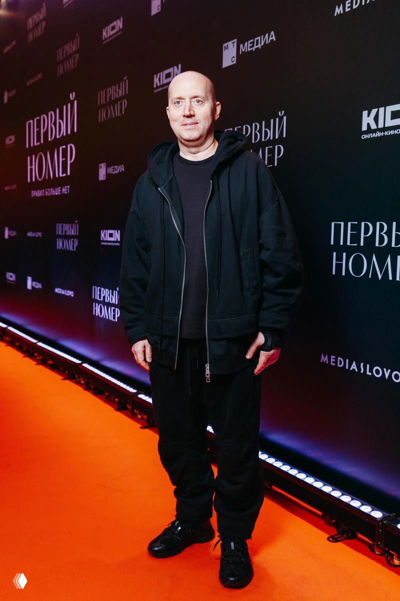 Мужчина в черной повседневной одежде позирует перед темной фотостеной с логотипами фильма; кадр с красной дорожки и промо-ивента.