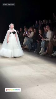 Девочка в пышном белом платье дефилирует по подиуму на показе Московской недели моды; в первом ряду зрители наблюдают и аплодируют, фото снято вертикально.