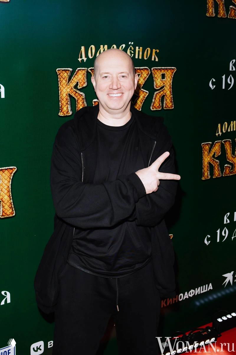 Мужчина в черной толстовке улыбается и позирует на фоне зеленой фотостены с названием проекта; портретный промо-кадр с мероприятия.