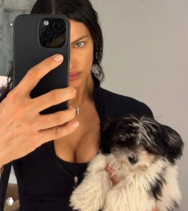 Крупный план: женщина делает селфи с телефоном, частично закрыв лицо, и держит пушистого щенка у груди на нейтральном фоне