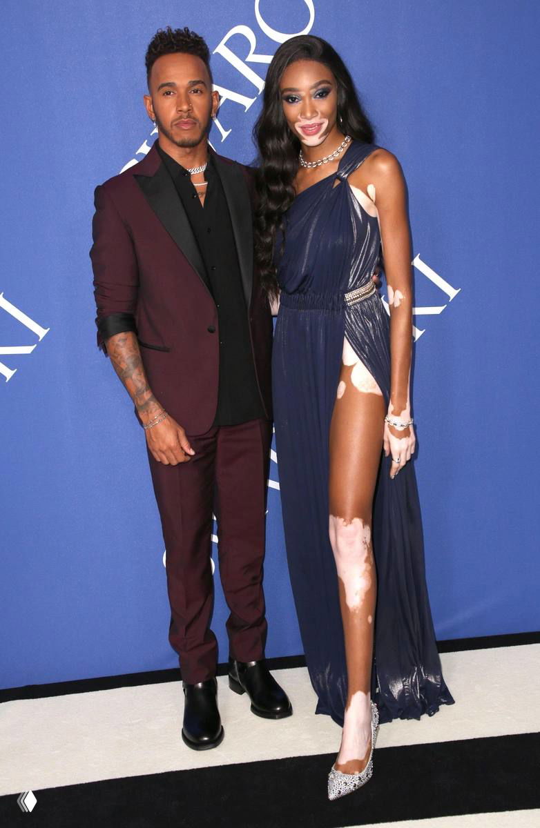 Мужчина в темном костюме рядом с моделью в длинном платье на синем фоне — официальный светский снимок на мероприятии, позирование для камер.