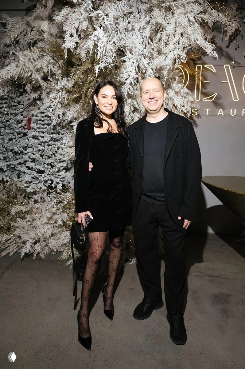 Пара на фоне украшенной ели фотозоны: мужчина в темной одежде рядом с женщиной в черном платье, совместный кадр с мероприятия и фотоколла.