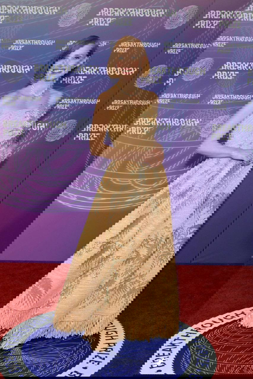 Женщина в бежевом объемном вечернем платье с вышивкой и бахромой позирует на красной дорожке на фоне баннера премии.