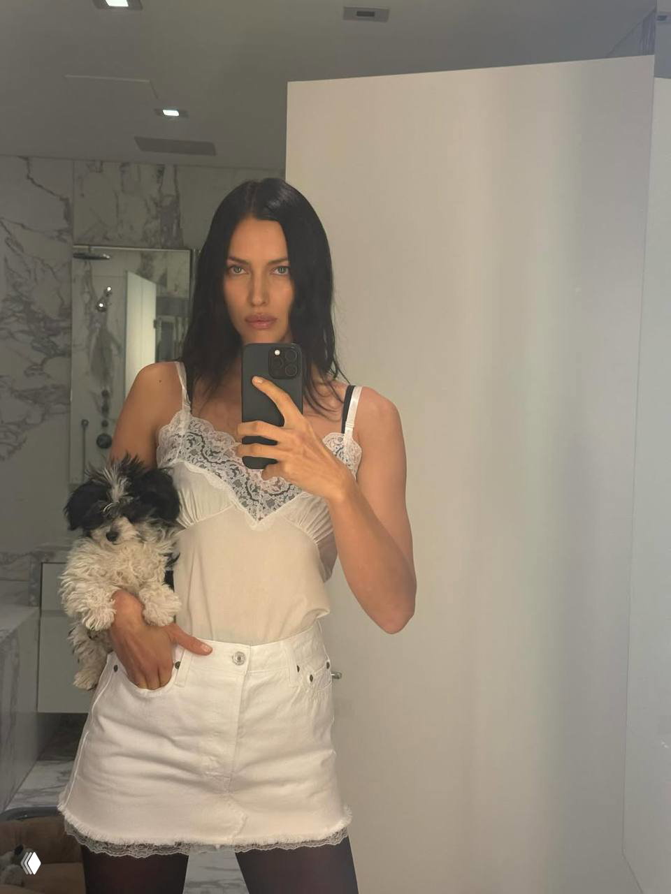 Женщина в белой лёгкой блузе и мини‑юбке стоит у зеркала и держит щенка на руке — интерьер ванной комнаты виден позади