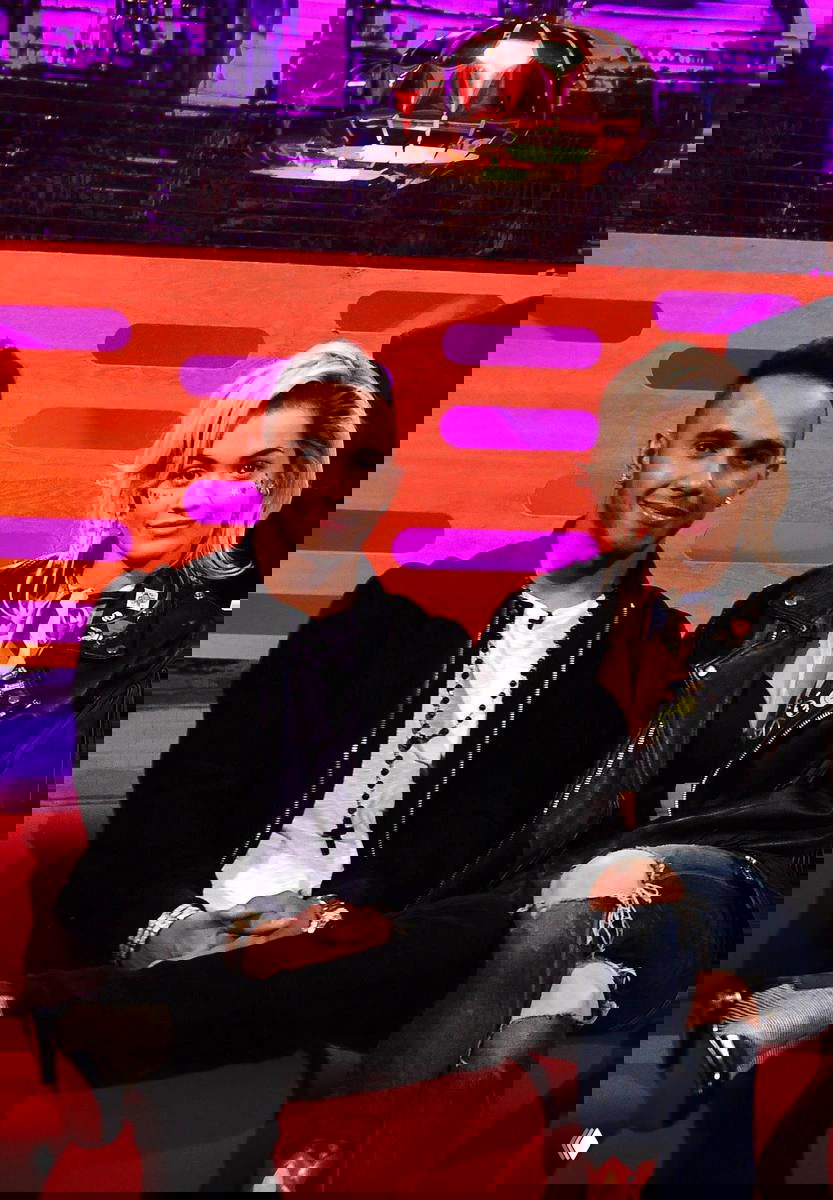 Мужчина и женщина сидят на телешоу на диване; интерьер студии ярко освещен, кадр в полудетали, элементы шоу и публика видны вблизи.