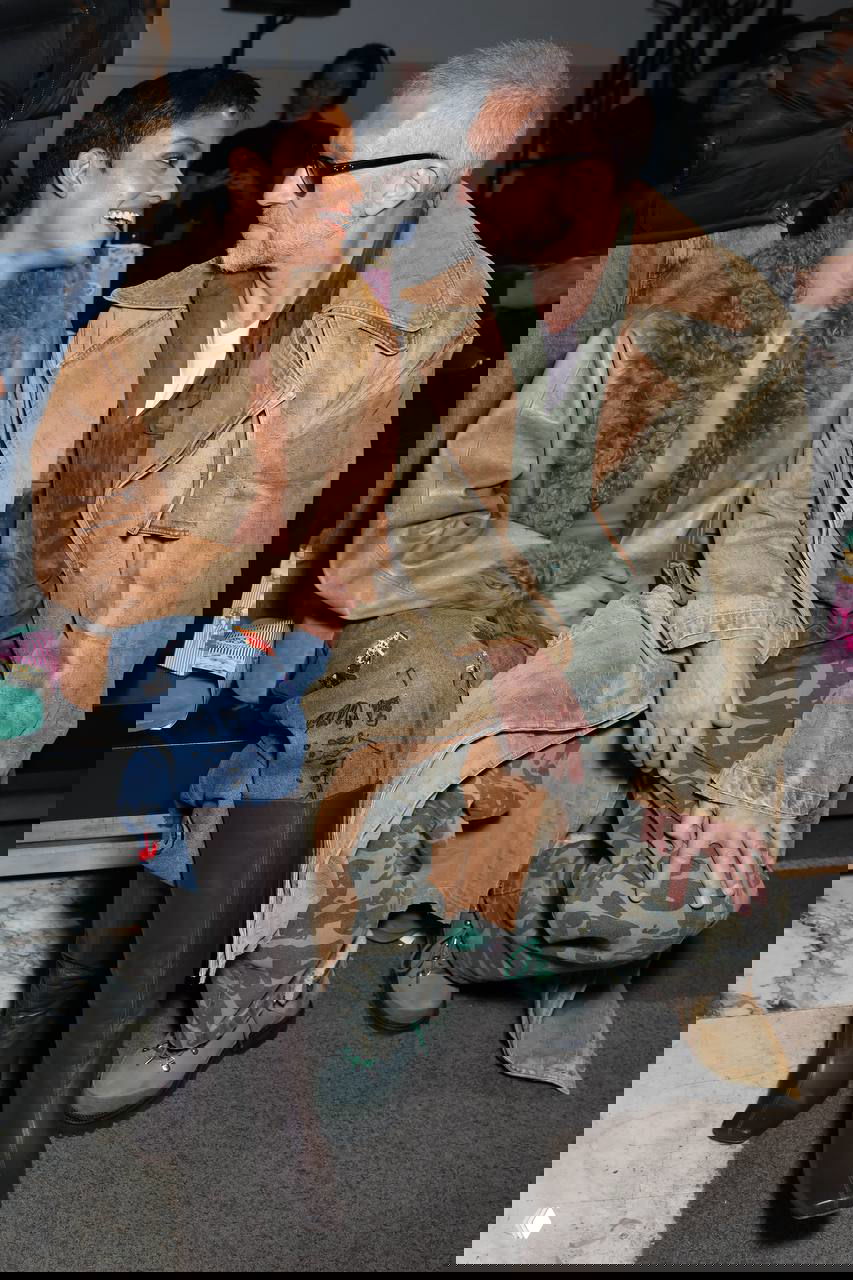 Пара сидит на фронтроу и беседует: женщина улыбается мужчине в неформальном образе на фоне зрительного зала модного показа.