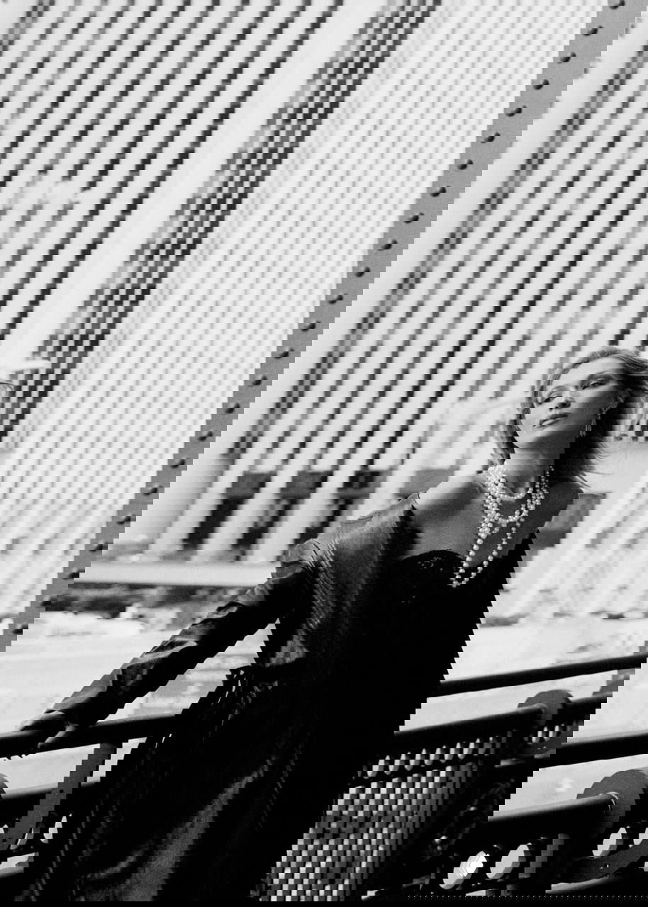 Чёрно-белая уличная съёмка модели с длинными жемчужными бусами на фоне городских высоток; ветреный профиль и архитектурный фон.