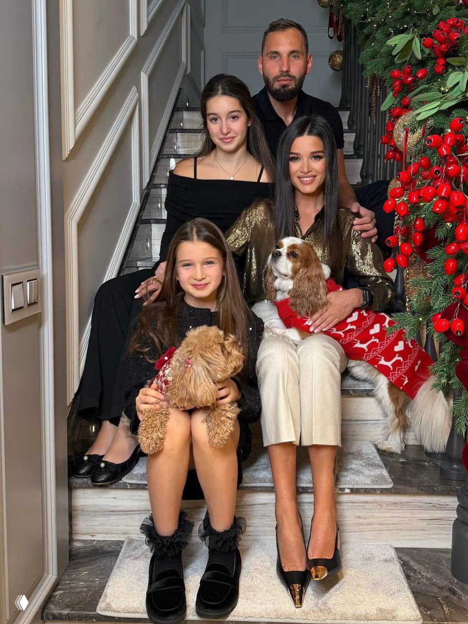 Портретная семейная сцена на ступеньках у украшенной гирляндами лестницы и ёлки: взрослые и дети держат маленьких собак в руках.