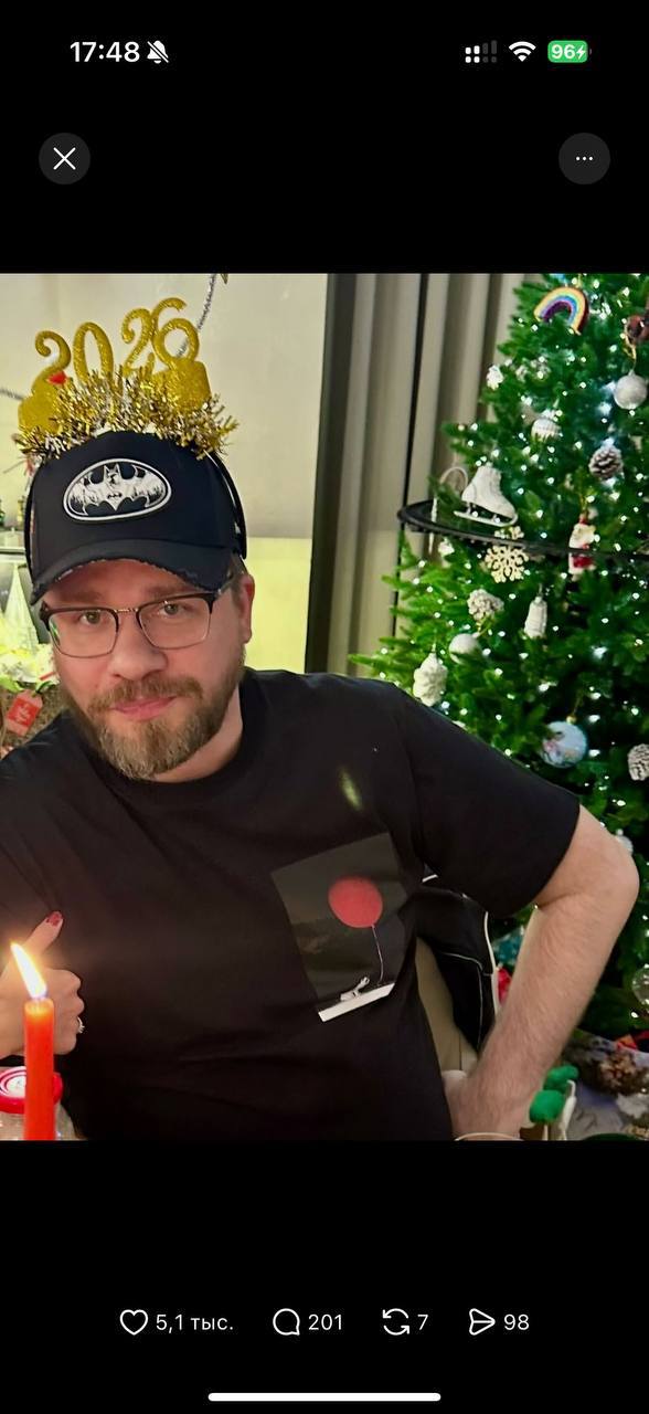Современное фото Гарика Харламова за праздничным столом в шапке с цифрой «2026», на фоне украшенная новогодняя ёлка и свеча.