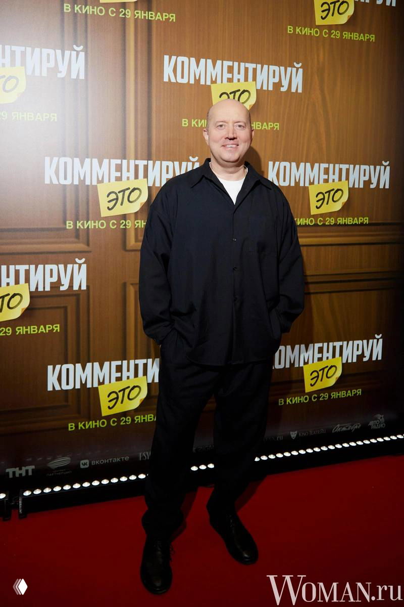 Мужчина в темной рубашке позирует на фоне постера с названием фильма «Комментируй это», кадр с красной дорожки и кинопоказа.