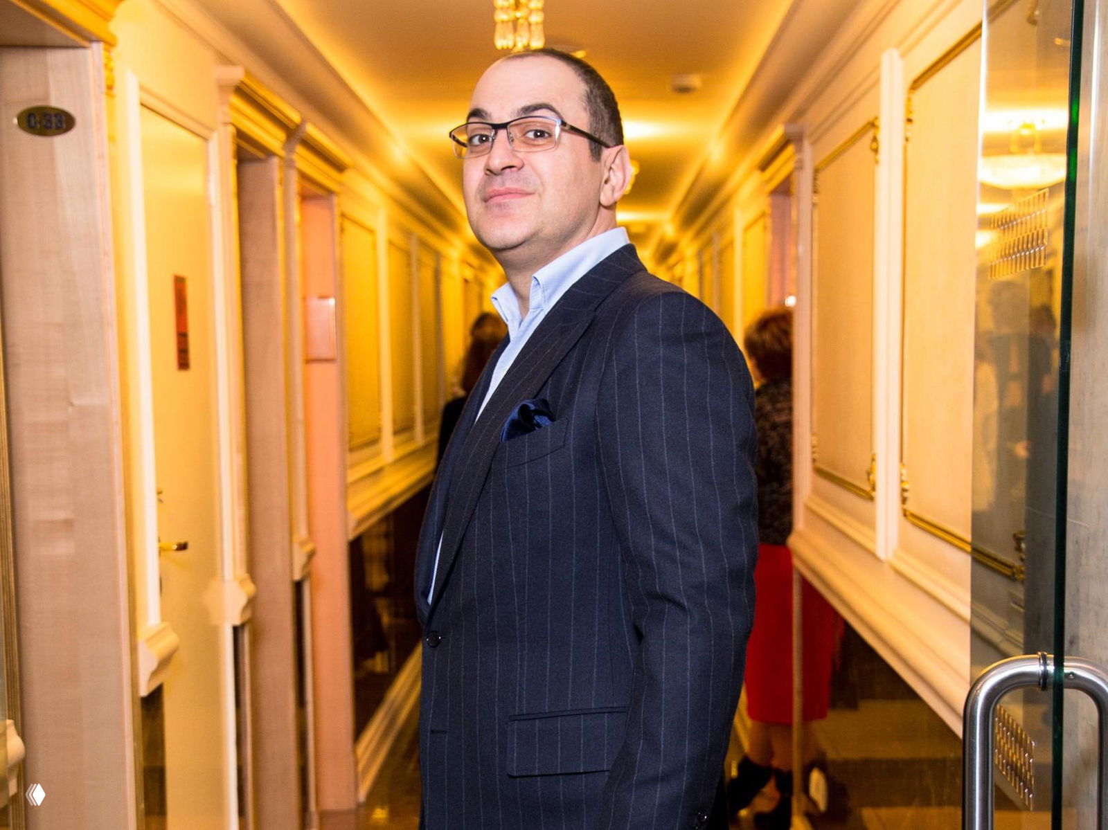 Фотография мужчины в тёмном полосатом костюме и очках в коридоре гостиницы с тёплым желтым освещением, кадр в полный рост.