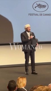 Джон Траволта на сцене Каннского фестиваля держит речь при вручении награды, кадр показывает его в полный рост на фоне экрана и зрителей.
