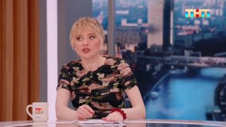 Женщина в теле-студии сидит за столом во время интервью; в кадре видны большие окна и городской пейзаж за ними.
