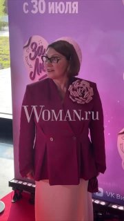 Женщина в бордовом жакете и белой юбке позирует на красной дорожке премьеры перед баннером с надписью «За любовь».