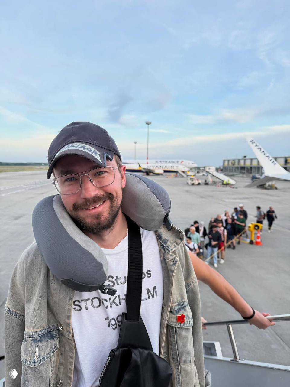 Александр Петров на посадочной полосе аэропорта с дорожной подушкой на шее, улыбается на фоне самолётов Ryanair и очереди пассажиров.