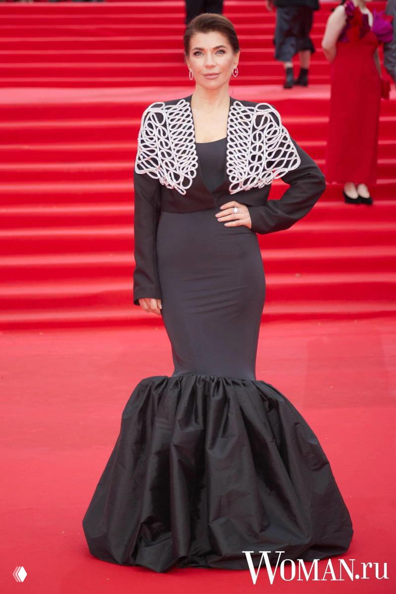 Женщина в приталенном платье-русалке темного оттенка с декоративным верхом позирует на красной дорожке кинофестиваля, полный рост.