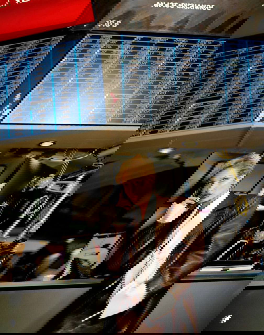 Модель в кожаном пальто в офисной обстановке: держит телефон, на заднем плане мониторы и торговые табло — деловой модный сет.