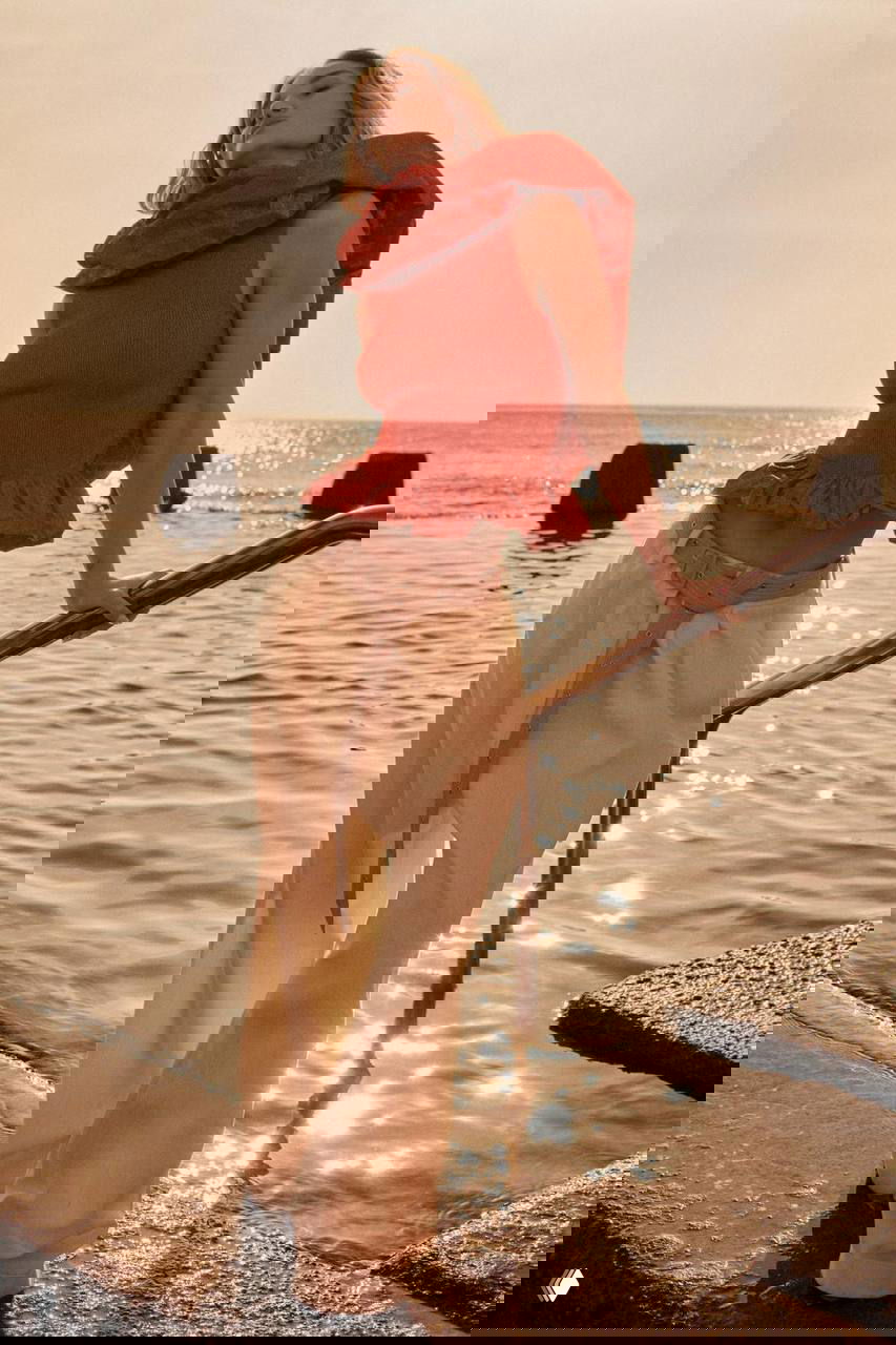 Роузи в красном топе у моря: фотосессия на пирсе при закате с расслабленной позой и солнцем в кадре.