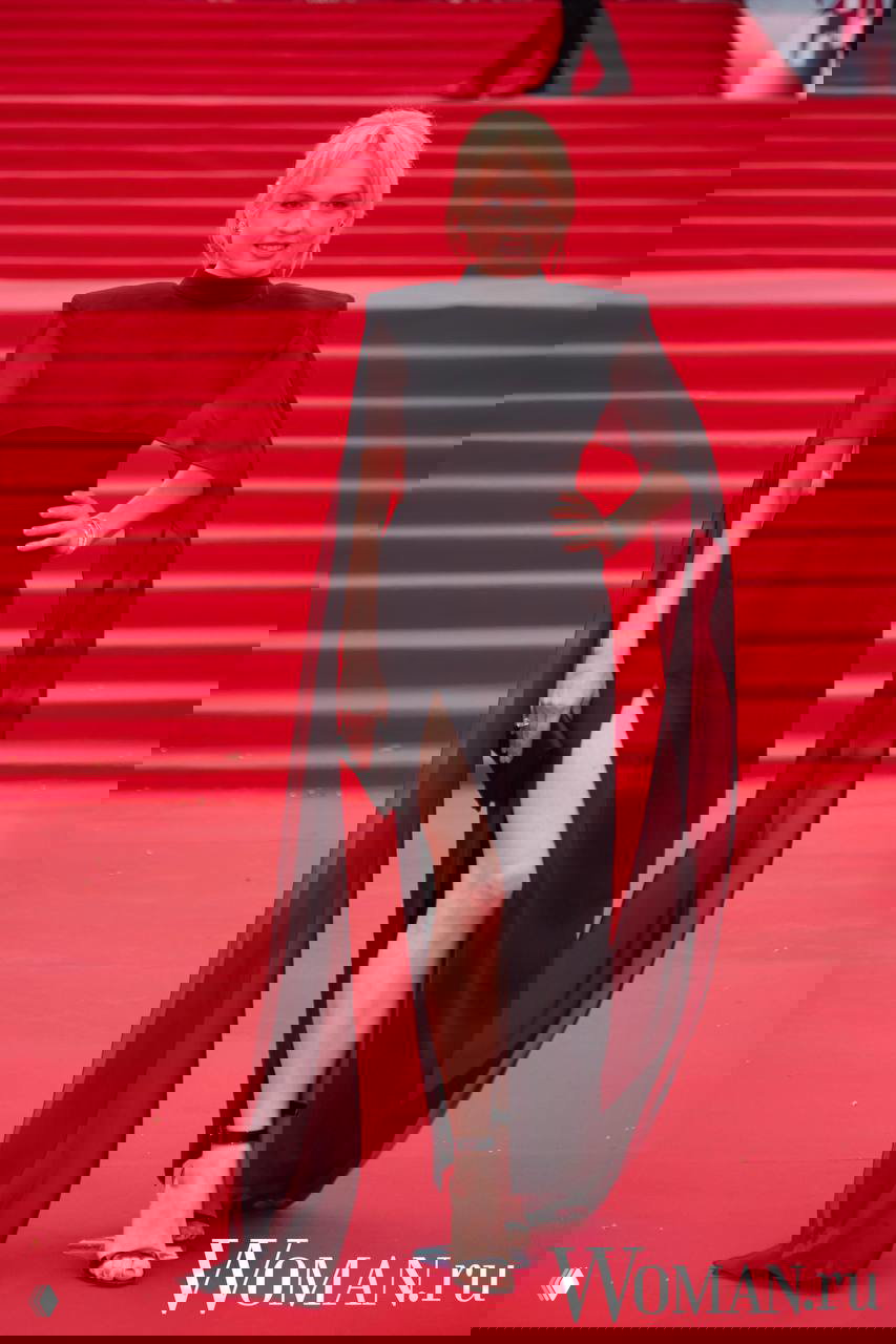 Женщина в черном платье с полупрозрачными рукавами и высоким разрезом идет по красной дорожке, видно силуэт и ткань наряда.