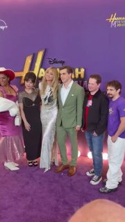 Вертикальный снимок с ковровой дорожки: группа гостей премьеры стоит у баннера «Hannah Montana» — мобильный ракурс со зрительного места.