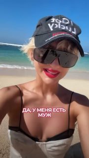 Крупный план женщины на пляже в солнцезащитных очках и кепке; на фото наложен текст «ДА, У МЕНЯ ЕСТЬ МУЖ», на фоне видны песок и море