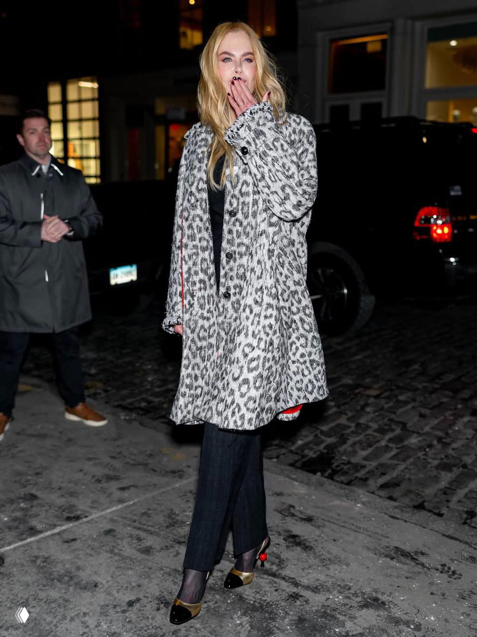 Фотография женщины в светлом пальто с принтом: поза на тротуаре, рука у лица, видно сочетание пальто с тёмными брюками и каблуками.