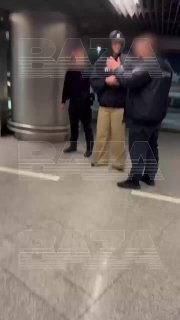 Размытое вертикальное видео в аэропорту: несколько мужчин в чёрной одежде у коридора, один держит телефон, кадр размытый.