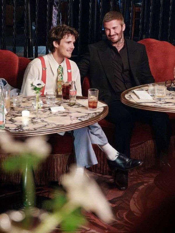 Двое мужчин в элегантной одежде сидят за мраморным столиком в ресторане, на столе напитки и декоративные свечи, ведут непринуждённую беседу.