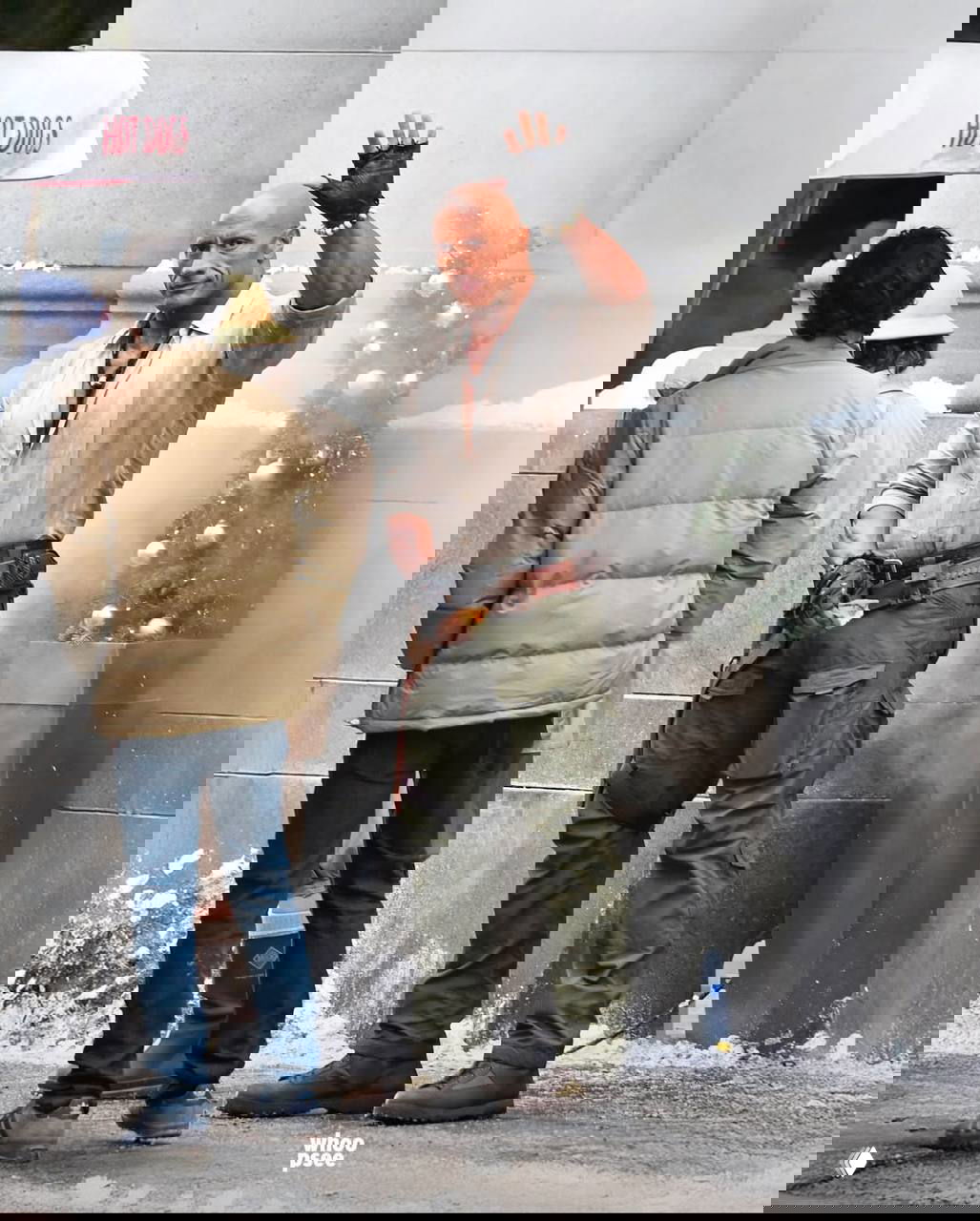 Мускулистый мужчина в рабочем костюме и перчатках машет рукой на съёмочной площадке; вокруг декорации и мокрый тротуар.